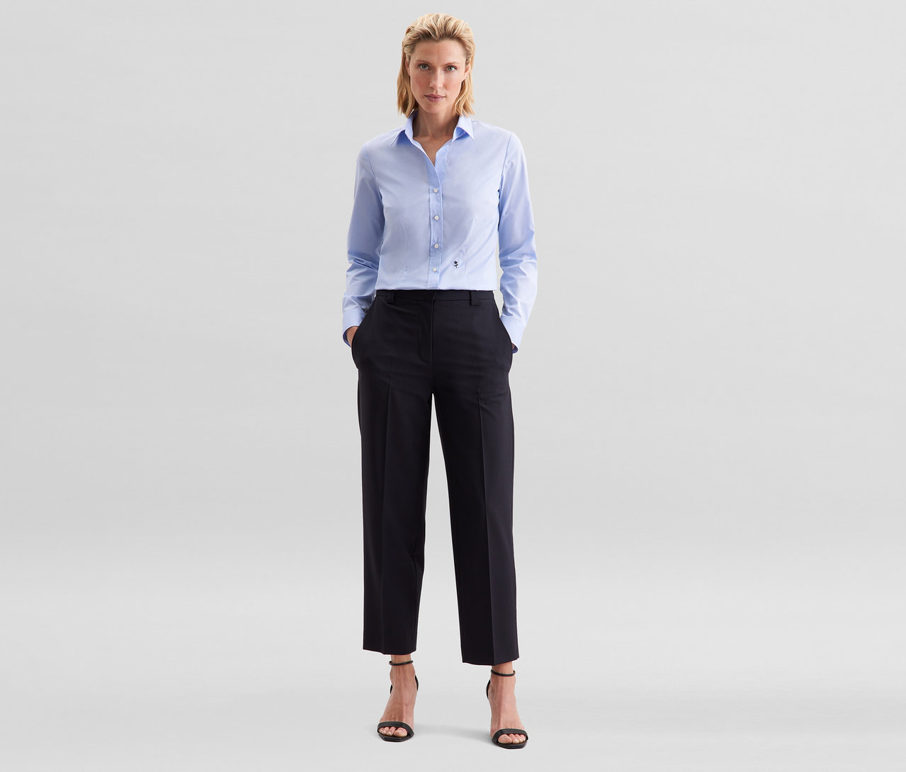 Žena stojí s rukami vo vreckách, má na sebe svetlomodrú blúzku Seidensticker Slim Fit non iron, čierne nohavice a čierne sandále na vysokom opätku.