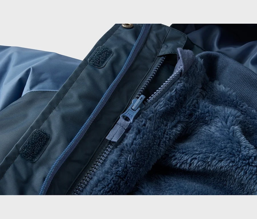 Detail modrej bundy, zobrazujúci jej tmavomodrú vonkajšiu látku, čiastočne rozopnutý zips a nadýchanú modrú flísovú podšívku. Na lége zipsu sú viditeľné suché zipsy.