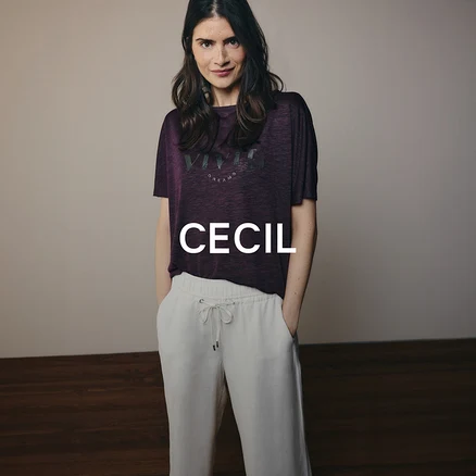 Žena s tmavými vlasmi má na sebe fialové tričko s nápisom a svetlé nohavice so šnúrkou. Stojí s rukami vo vreckách a pozerá sa do kamery.