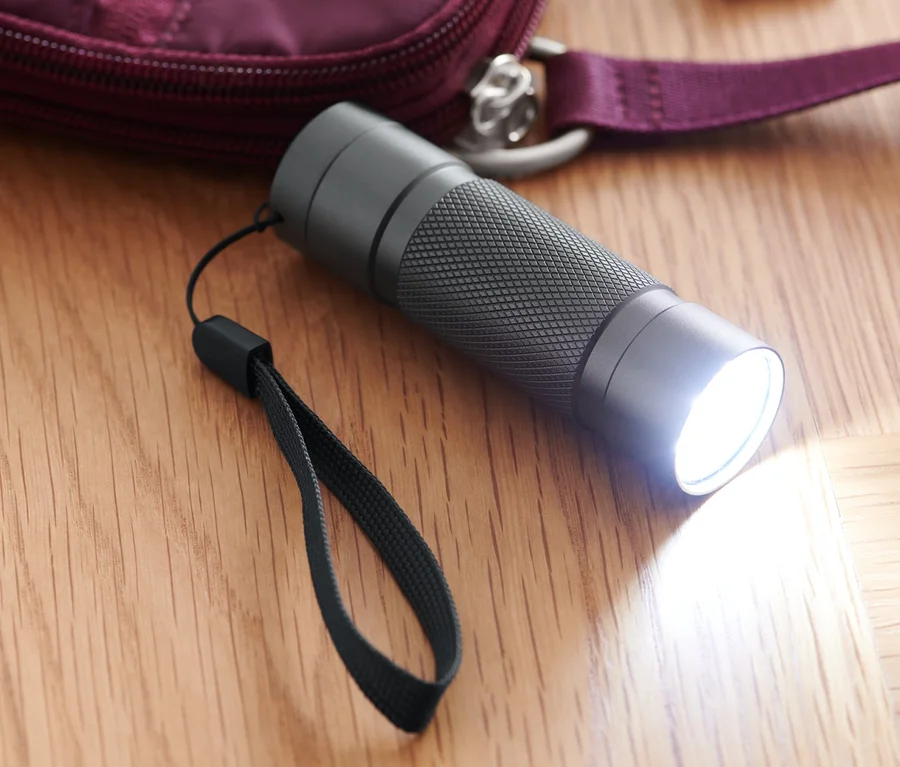 Svietiaca vrecková lampa s LED leží na drevenej podlahe vedľa bordovej kabelky a kľúčov.