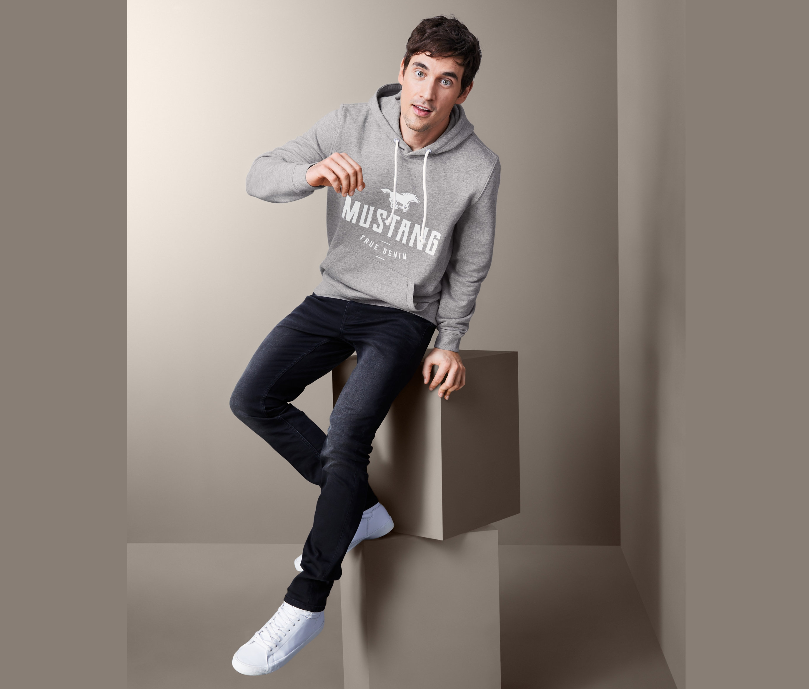 Muž sedí na kocke a má na sebe sivú mikinu s kapucňou Mustang, džínsy a tenisky.