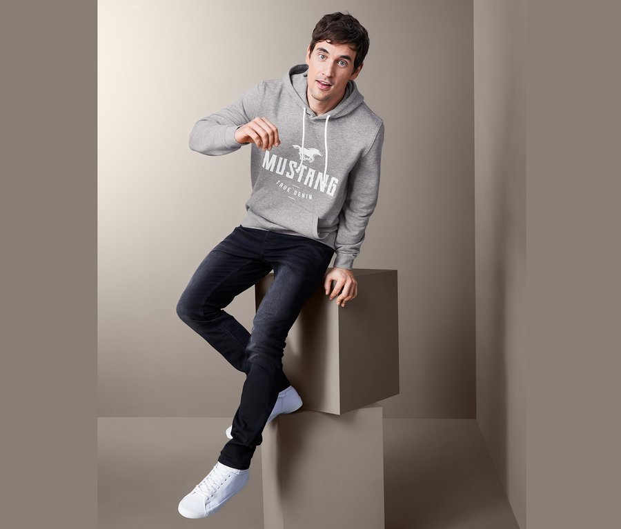 Muž sedí na kocke a má na sebe sivú mikinu s kapucňou Mustang, džínsy a tenisky.