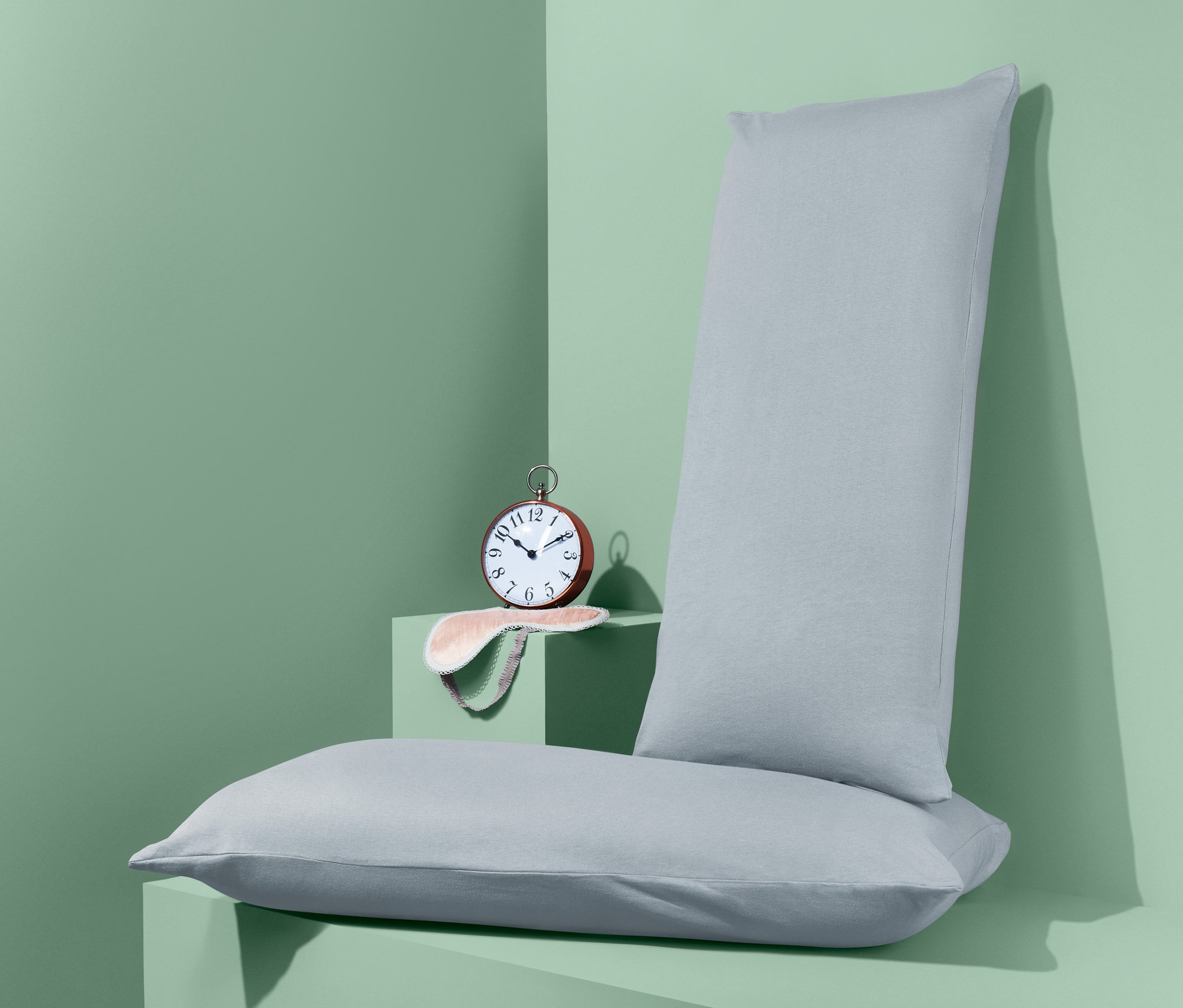 Dve džersejové obliečky na vankúše ležia na sebe na zelenom povrchu vedľa budíka a masky na spanie.