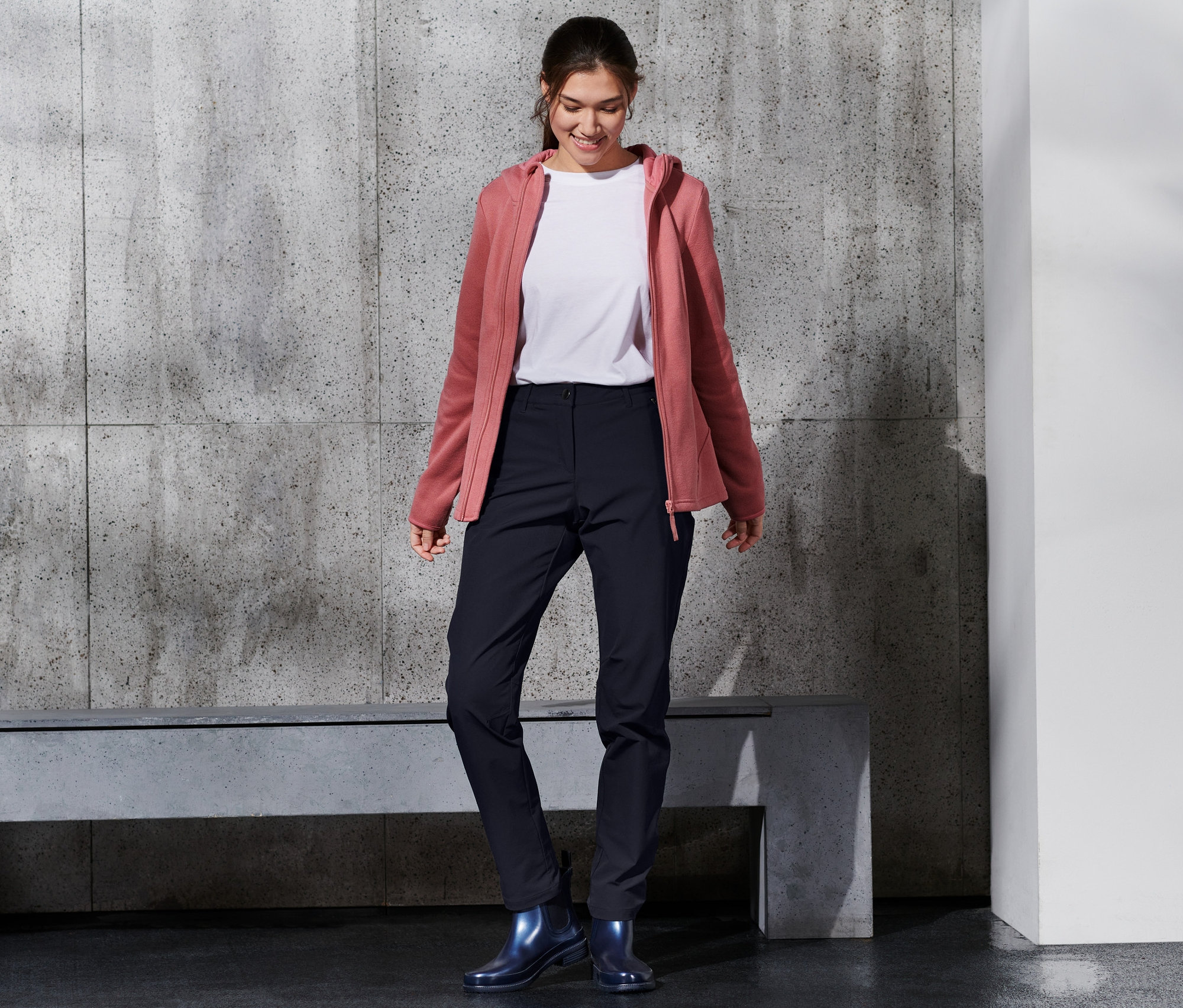 Usmievajúca sa žena má na sebe softshellové nohavice, biele tričko a ružovú mikinu s kapucňou.