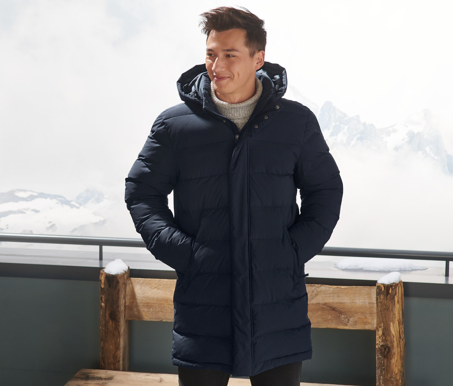 Muž v tmavomodrej páperovej prešívanej parke stojí vonku s rukami vo vreckách.