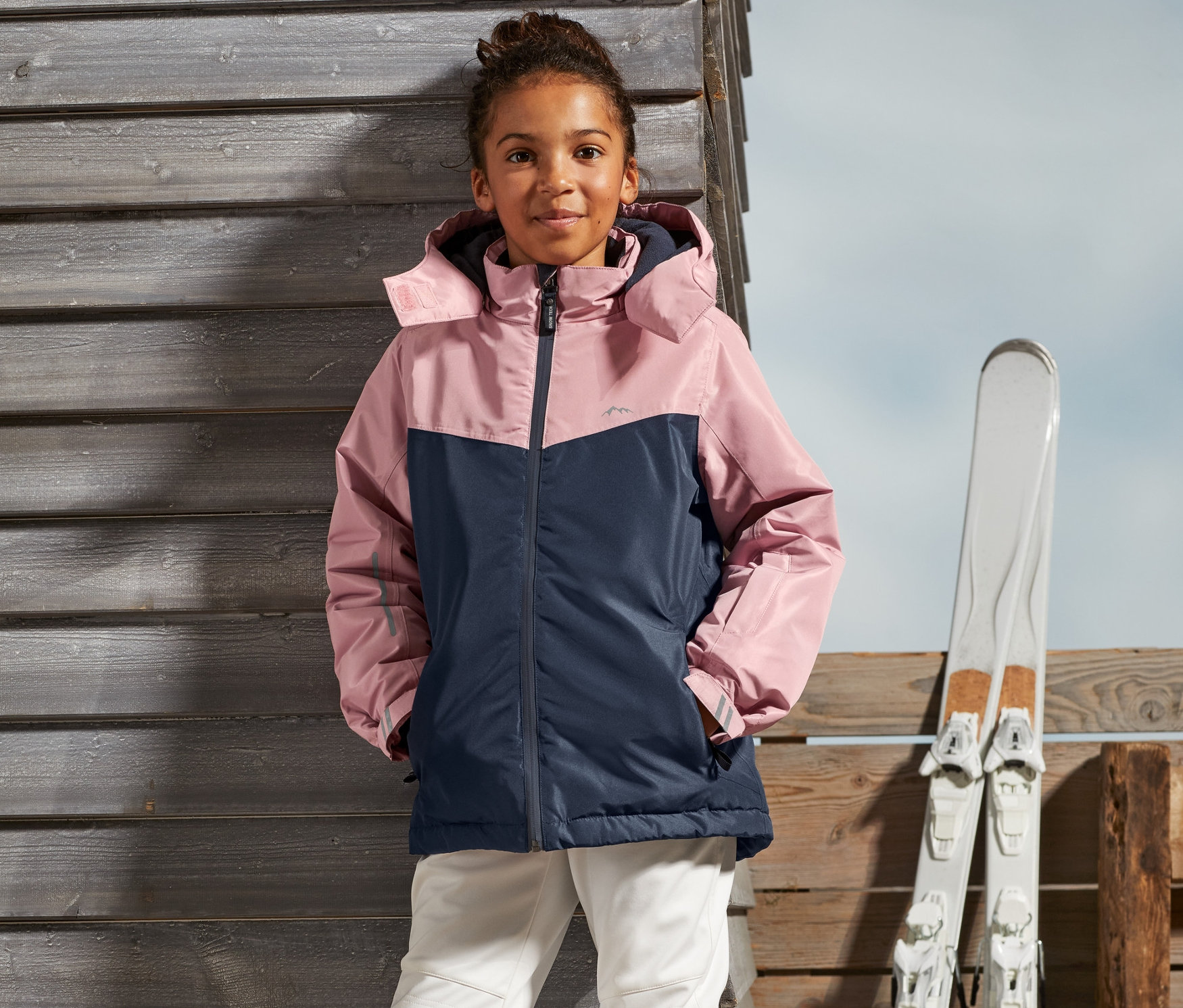 Dievča stojí vonku v dievčenskej lyžiarskej bunde a lyžiarskych nohaviciach.
