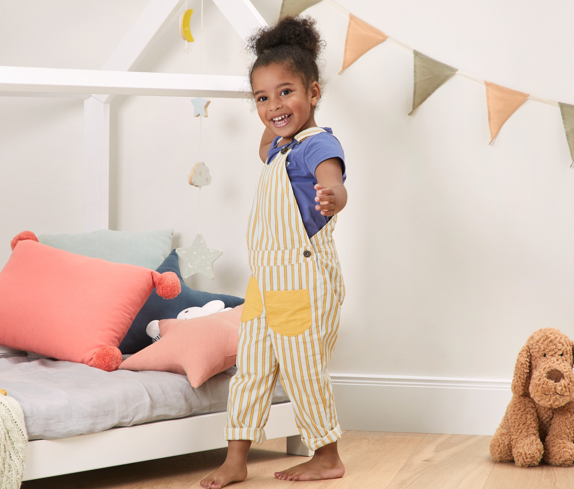 Usmievajúce sa dievčatko stojí v detskej izbe a má na sebe žlté pruhované nohavice s náprsenkou.