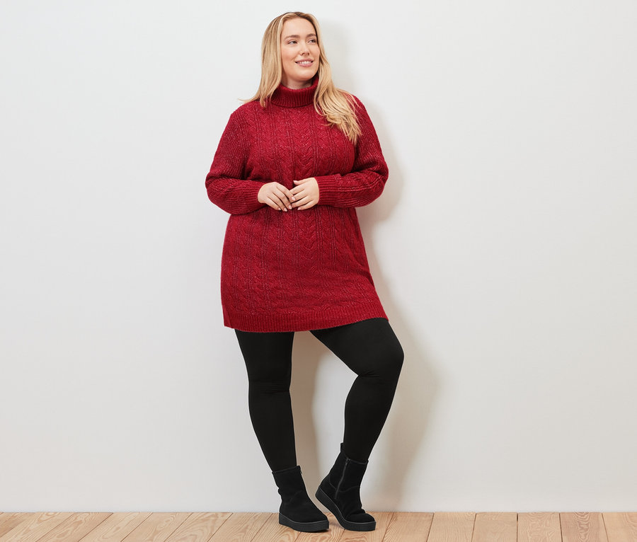 Žena pózuje v dlhom červenom pletenom pulóvri s vrkočovým vzorom, čiernych legínach a topánkach.