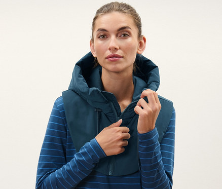 Žena si upravuje vatovanú kapucňu so stojatým golierom, má na sebe modré pruhované tričko a čierne nohavice.