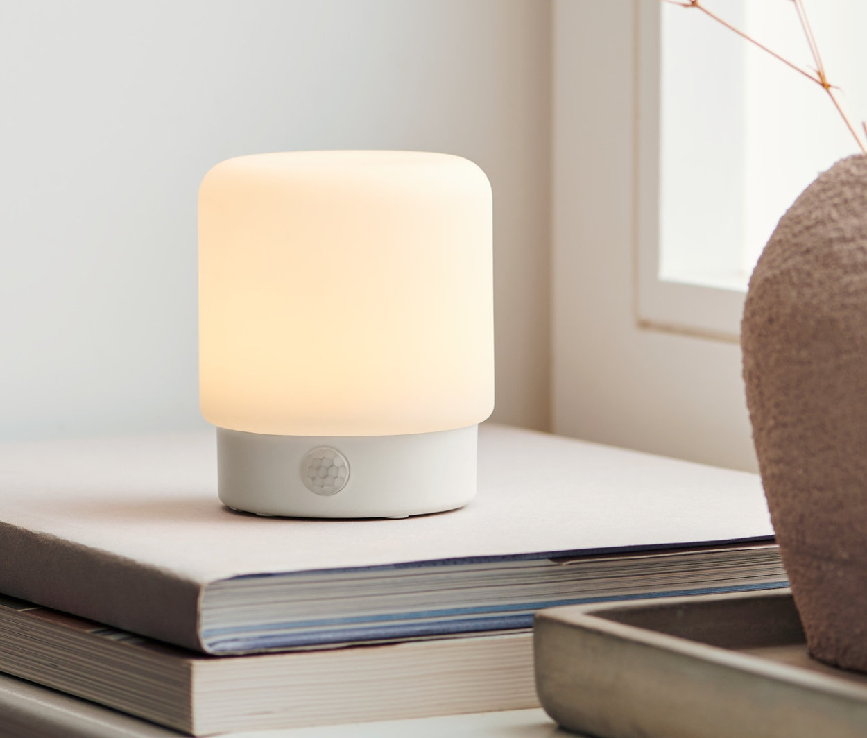 Malé multifunkčné svietidlo s LED diódami a so snímačom pohybu stojí na knihách vedľa vázy s kvetmi.