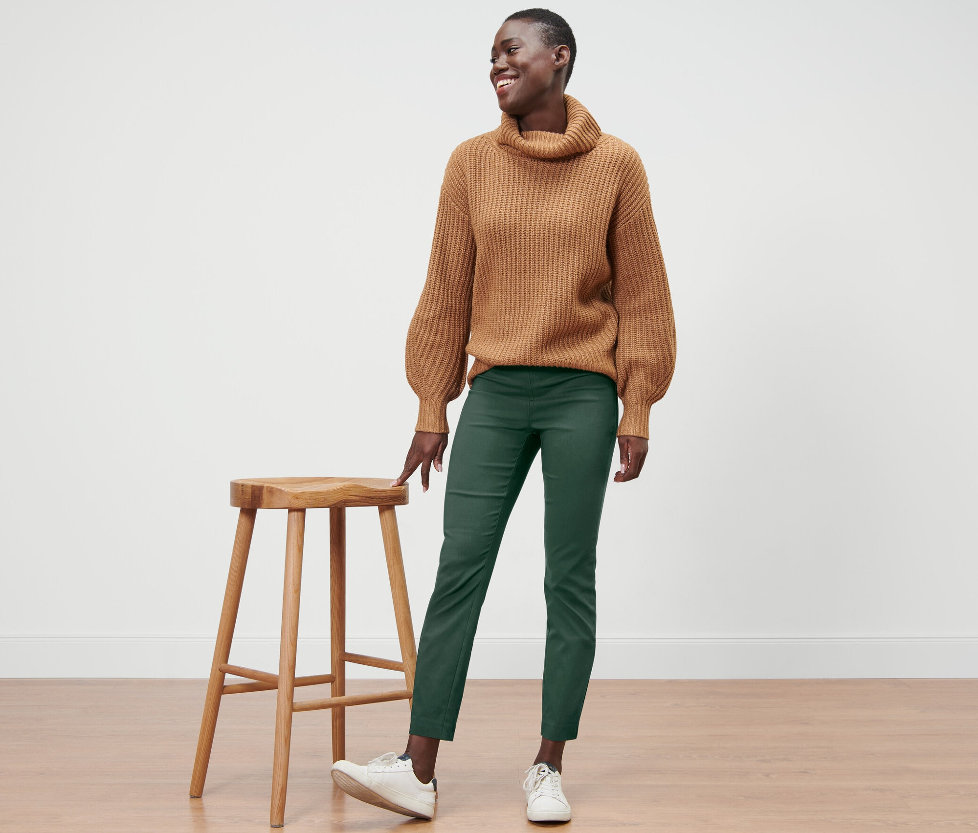 Usmievajúca sa žena stojí vedľa drevenej stoličky a má na sebe hnedý sveter a zelené strečové nohavice.