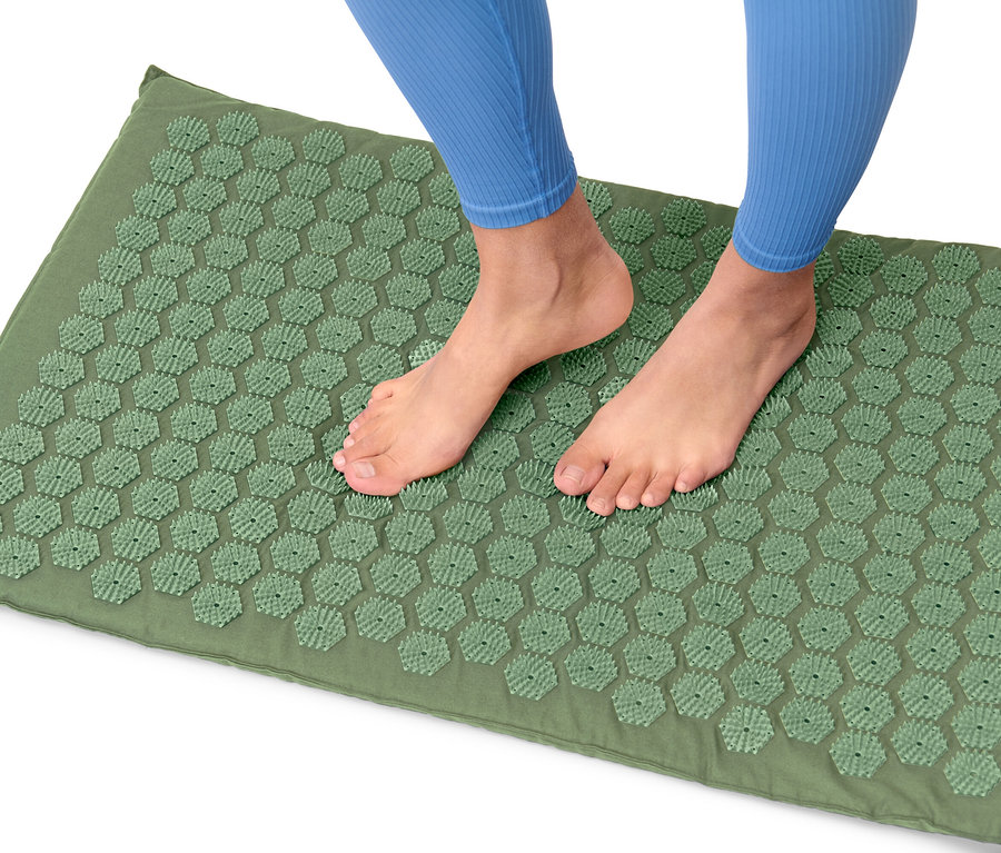 Nohy osoby v modrých legínach stoja na zelenej akupresúrnej podložke.