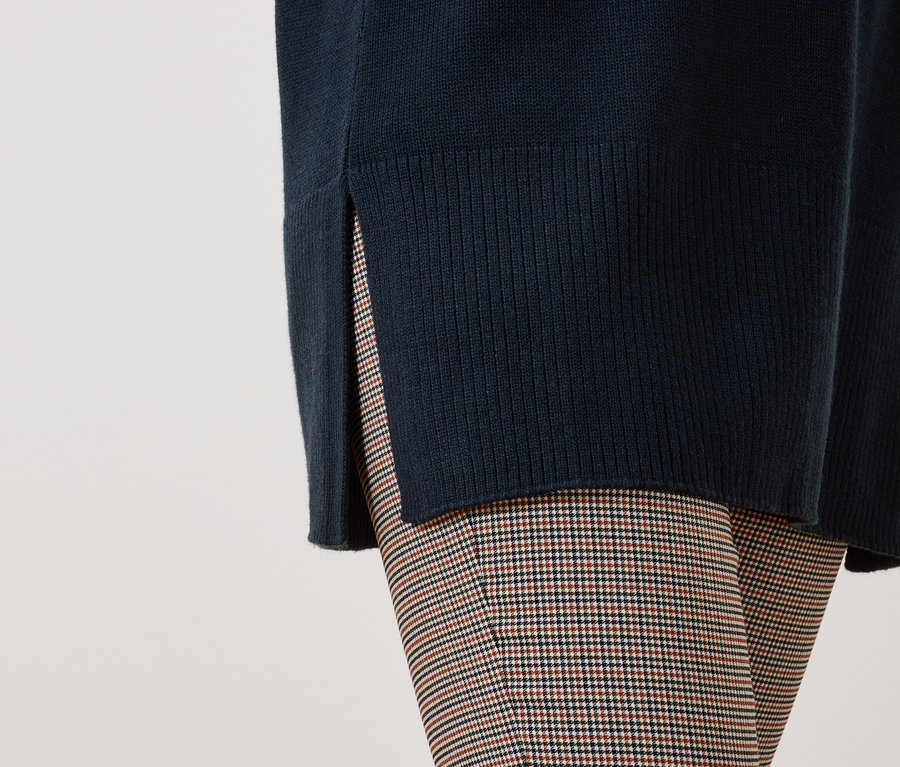Detail dámskeho outfitu s tmavomodrým svetrom s rázporkom a kockovanými nohavicami.