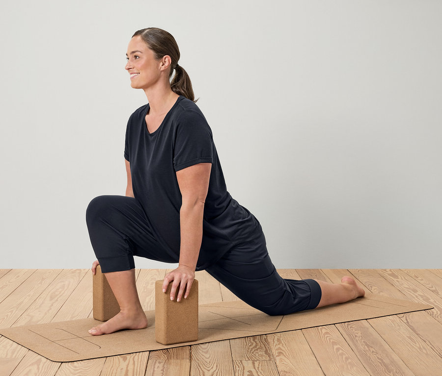 Žena v čiernom športovom oblečení robí výpad na hnedej podložke na jogu s blokmi.