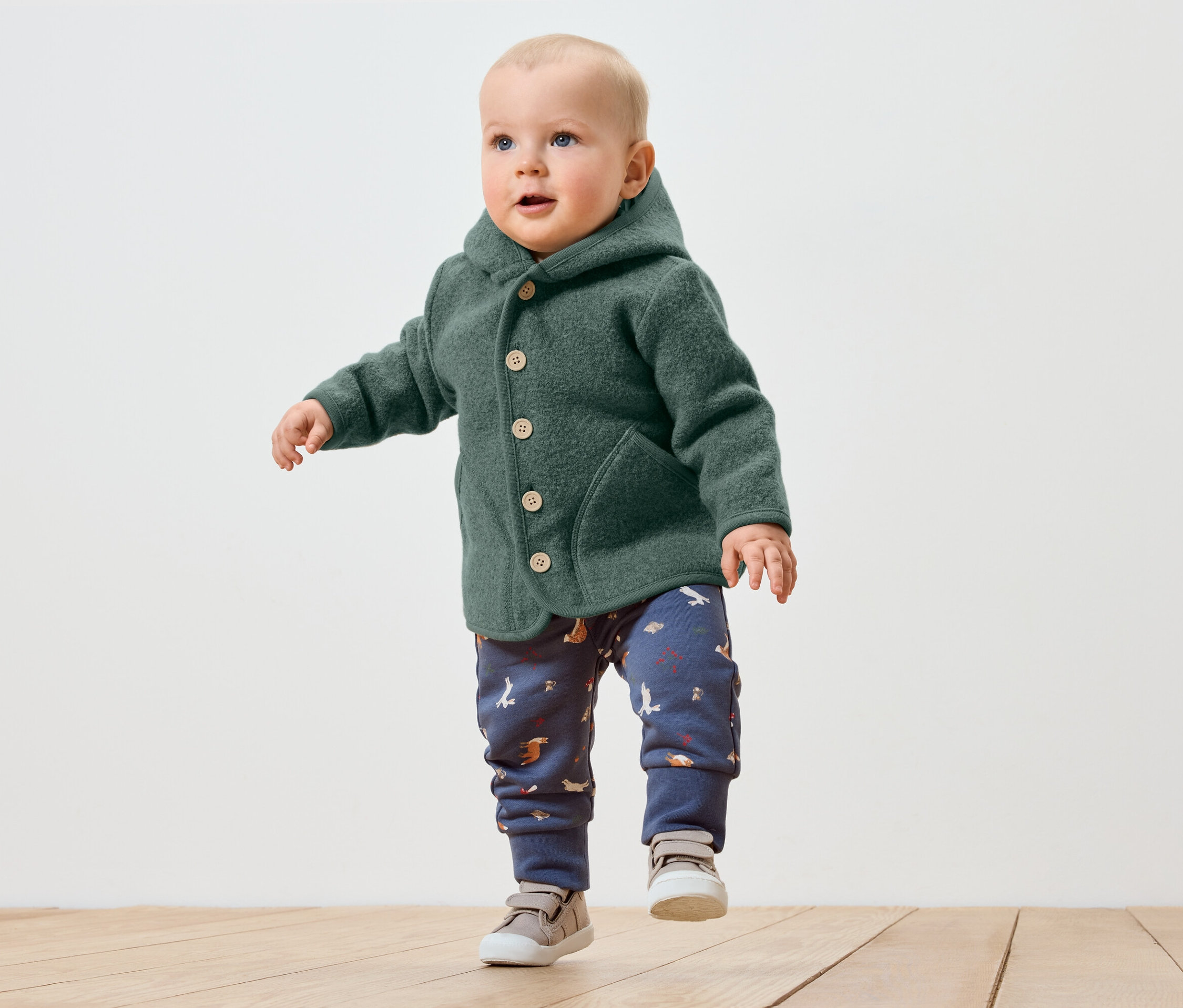 Batoľa v zelenej vlnenej bunde s kapucňou a vzorovaných nohaviciach robí prvé kroky po drevenej podlahe.