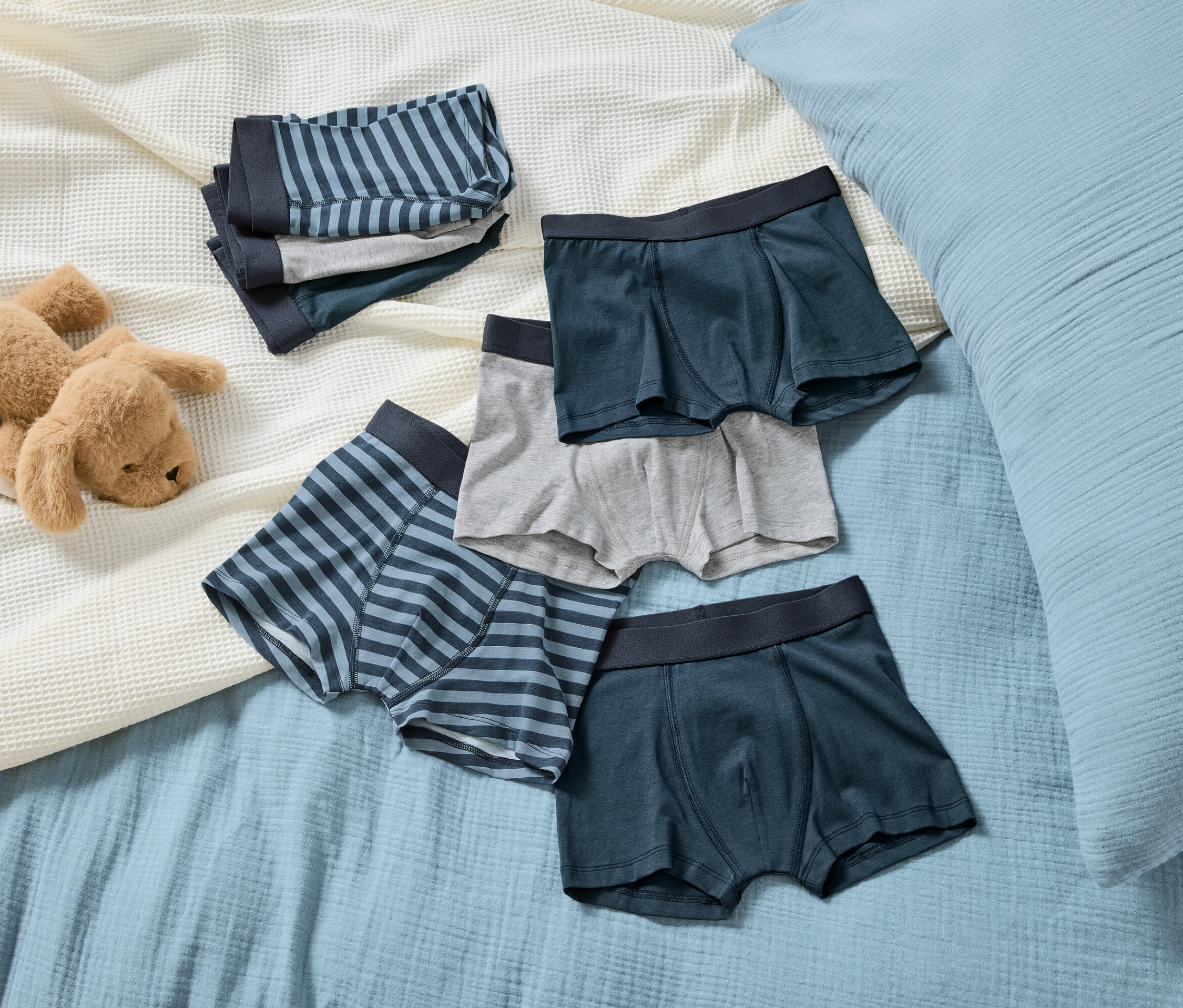 Na svetlomodrej deke leží hnedý plyšový pes vedľa kôpky pruhovaných a jednofarebných chlapčenských boxeriek.