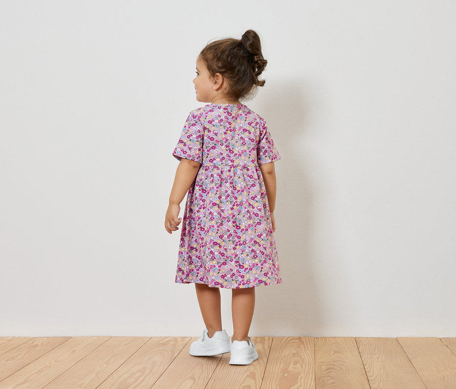 Dievčatko v kvetinových šatách a bielych teniskách stojí chrbtom ku kamere.