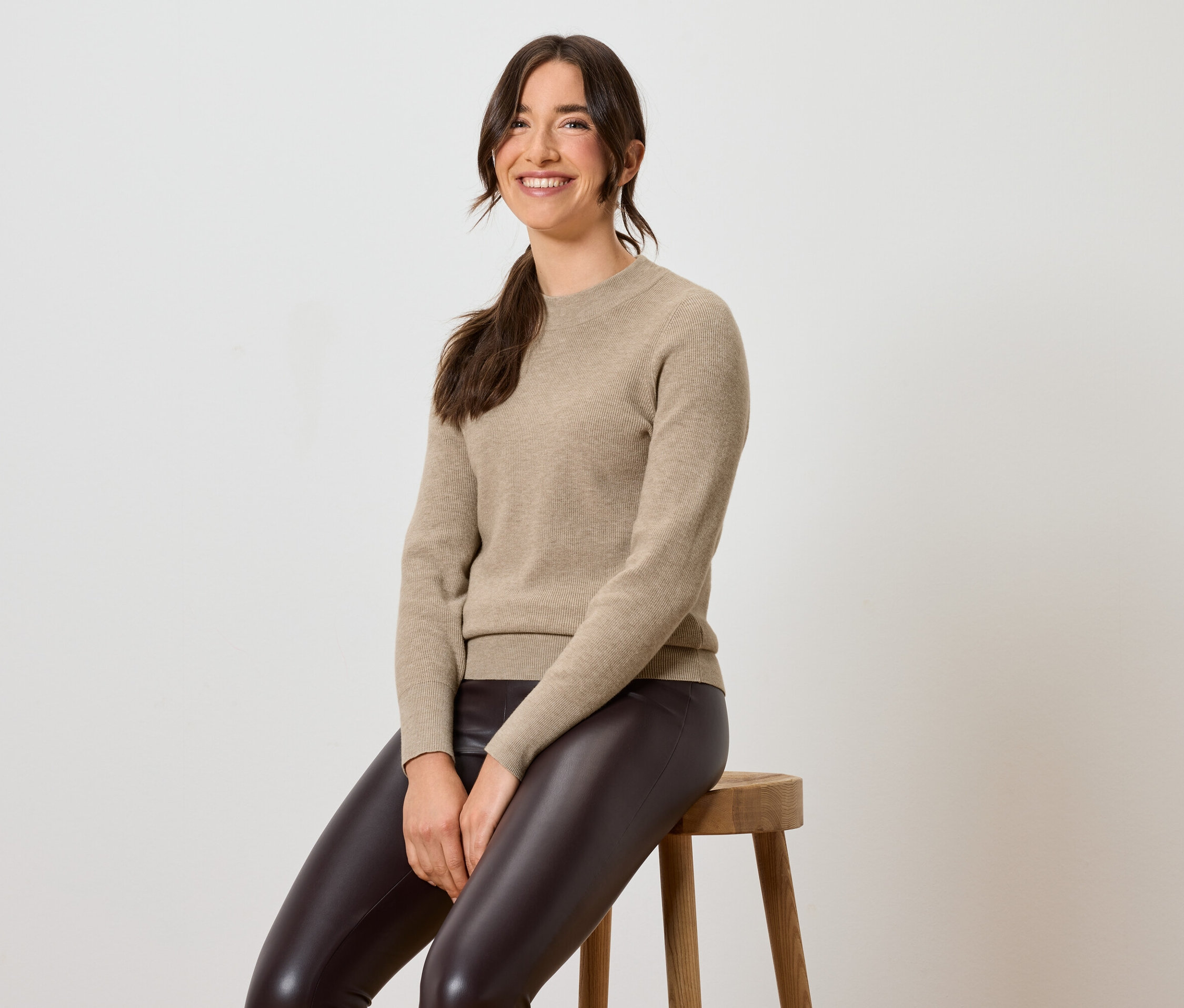 Žena sedí na stoličke v hnedých kožených legínach a béžovom svetri.