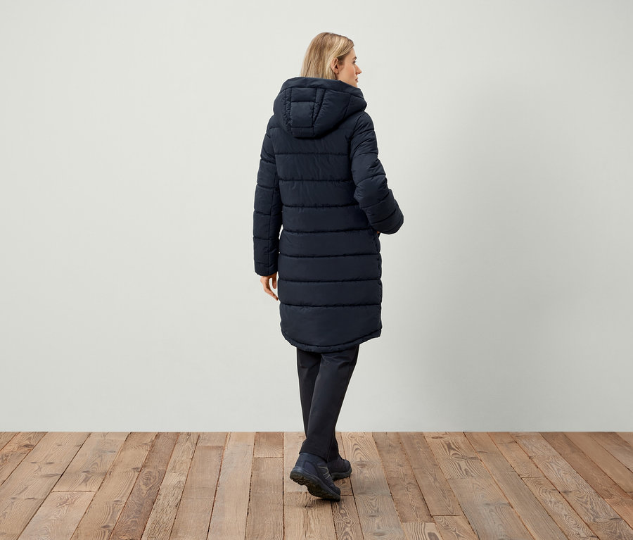 Žena v tmavomodrej prešívanej bunde s kapucňou, chrbtom k fotoaparátu.