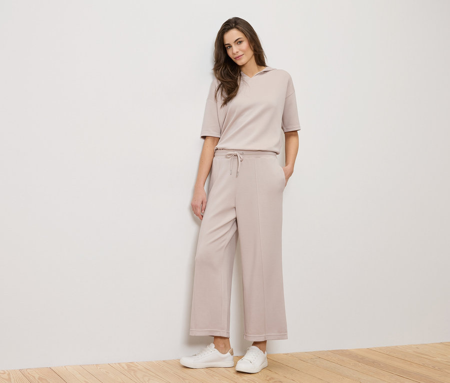 Žena stojí pred bielou stenou a má na sebe béžový outfit a biele tenisky.