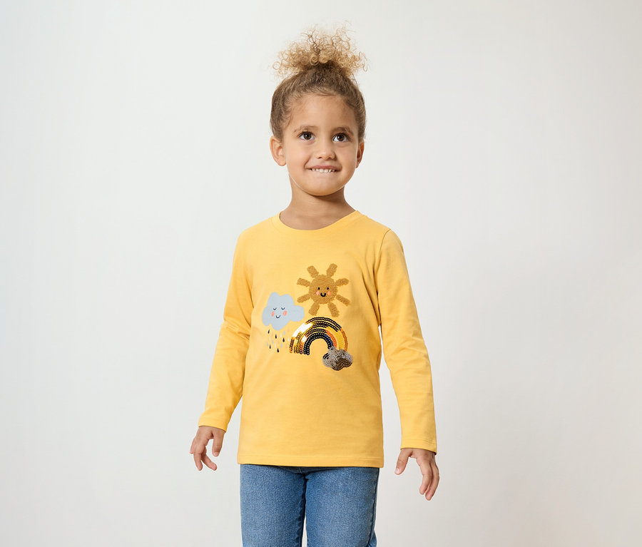 Dievča s kučeravými vlasmi má na sebe žlté tričko s dlhým rukávom s oblakom, slnkom a dúhou a modré džínsy.