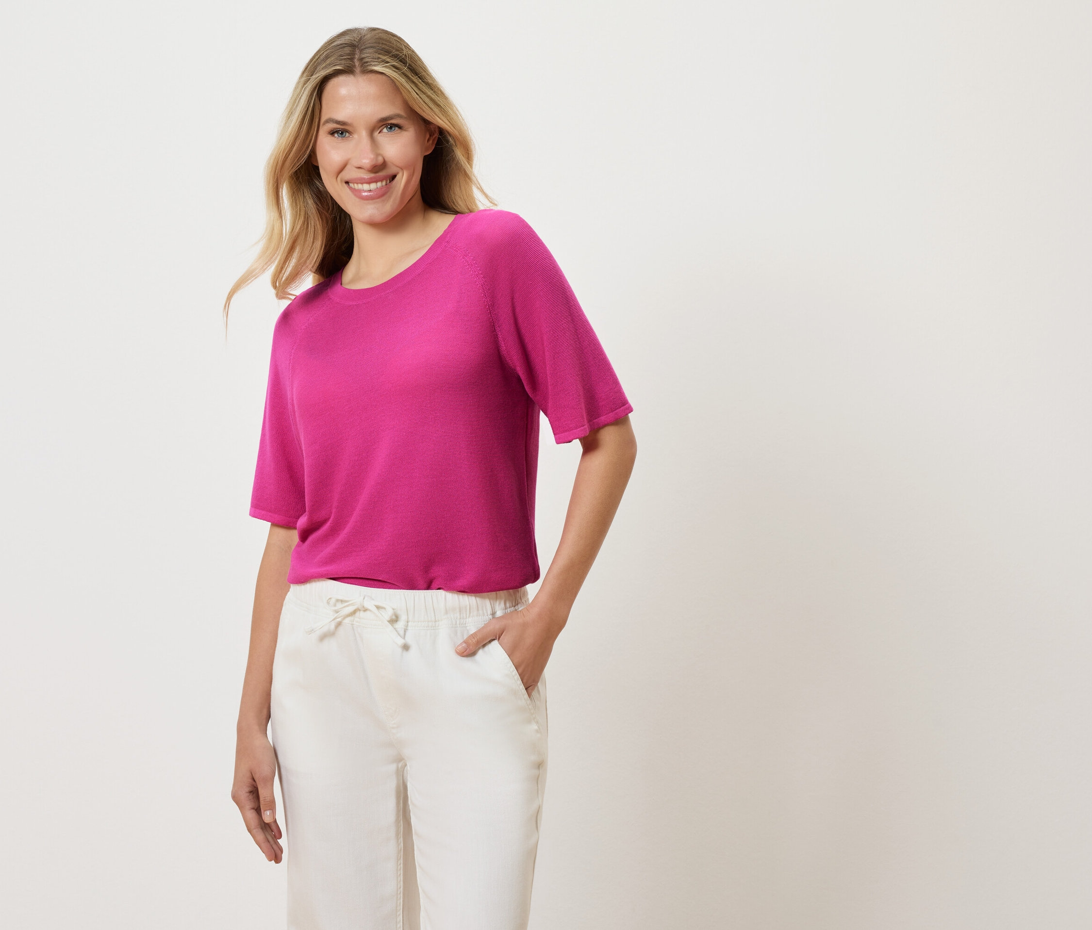 Žena s blond vlnitými vlasmi má na sebe ružové tričko a biele nohavice a dáva si ruku do vrecka.