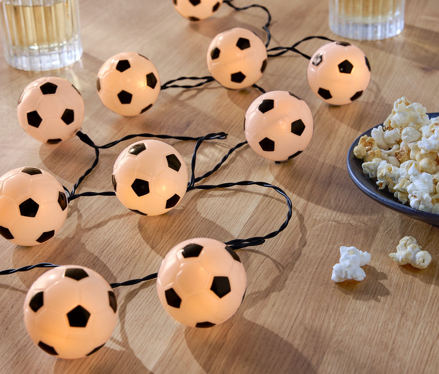 Svetelná reťaz v tvare futbalovej lopty leží na drevenom stole vedľa misky s popcornom a dvoch pohárov s nápojmi.