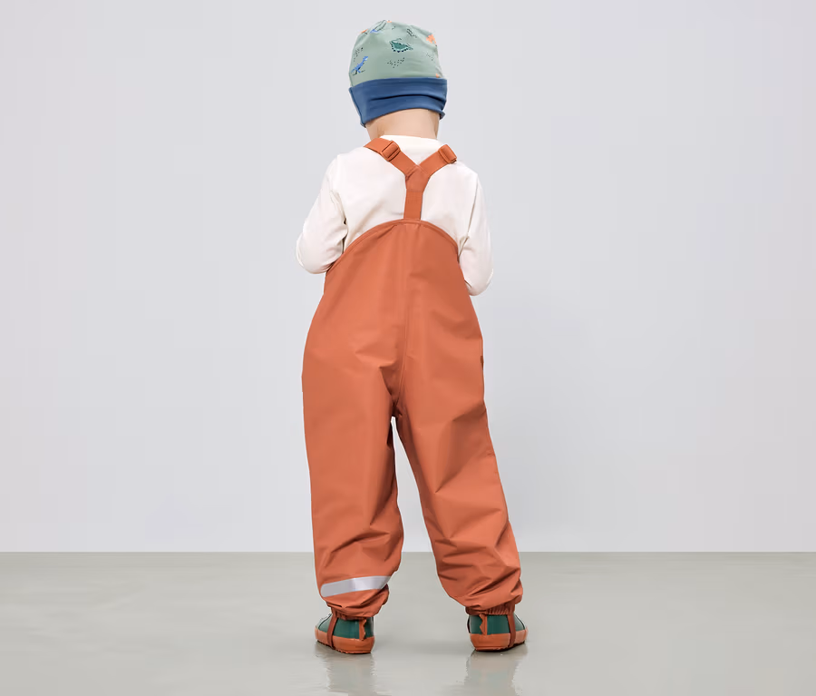Dieťa zozadu v oranžových nohaviciach s trakmi, zeleno-modrej čiapke a krémovom tope.
