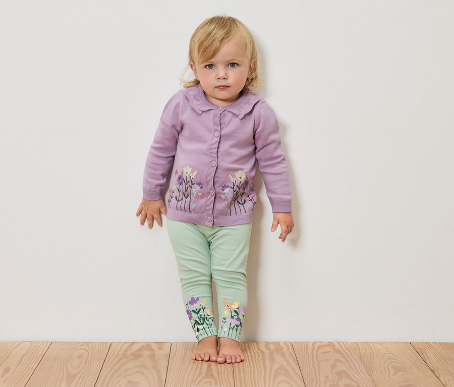 Blondína stojí naboso pred bielou stenou, má na sebe fialový pletený sveter s kvetinovým vzorom a zelené legíny s kvetmi na nohaviciach.