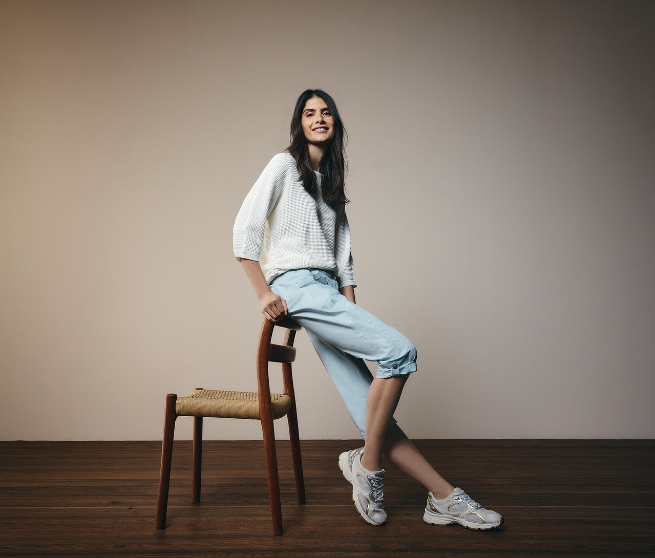 Žena sa opiera o stoličku, má na sebe svetlomodré 3/4 nohavice Cecil »New York« a biely sveter.