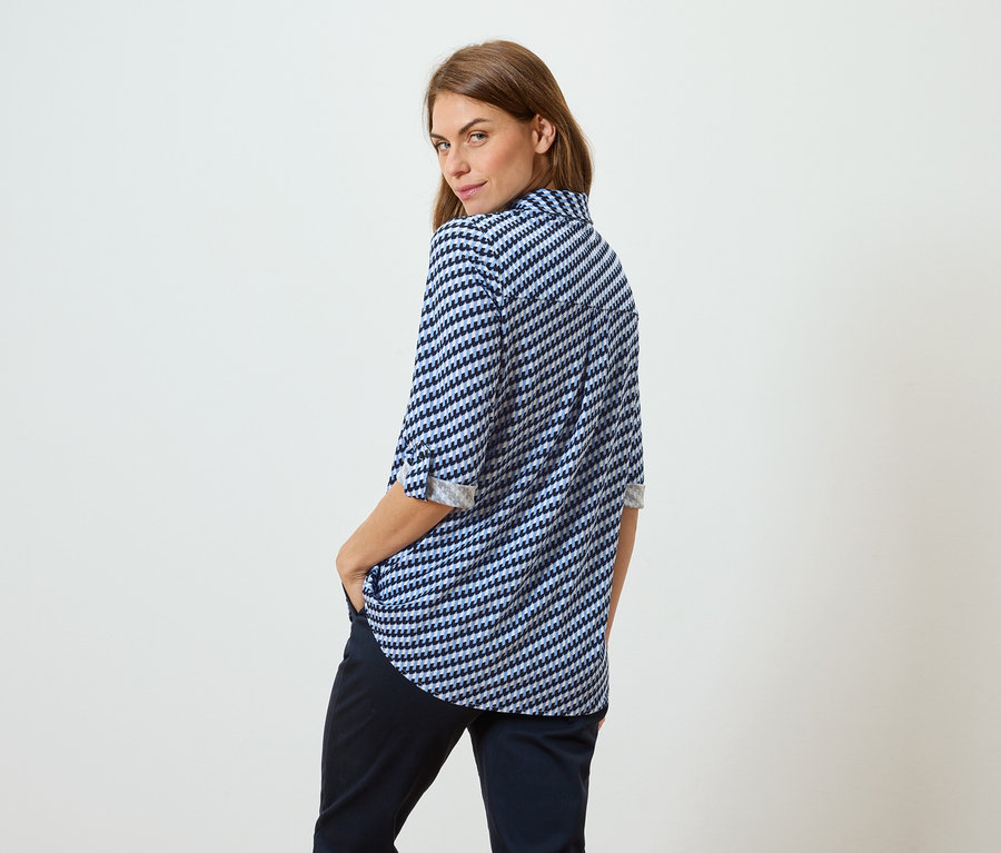 Žena s hnedými vlasmi v modro-bielej blúzke a tmavých nohaviciach stojí chrbtom k divákovi a pozerá cez plece.