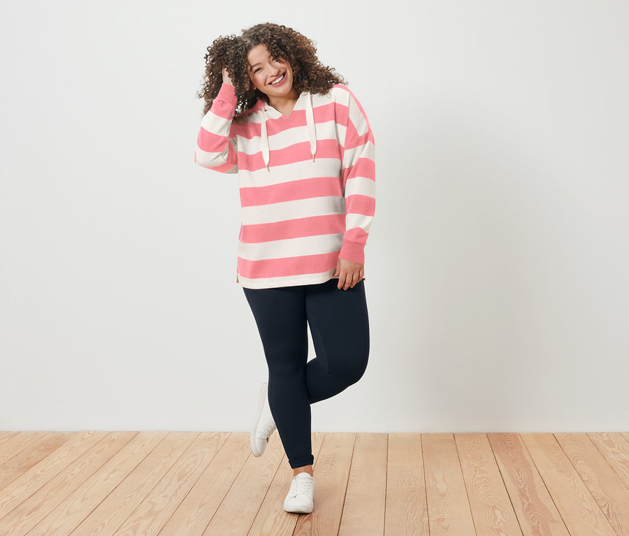 Žena s kučeravými vlasmi v pásikavej mikine s kapucňou a legínach sa usmieva.