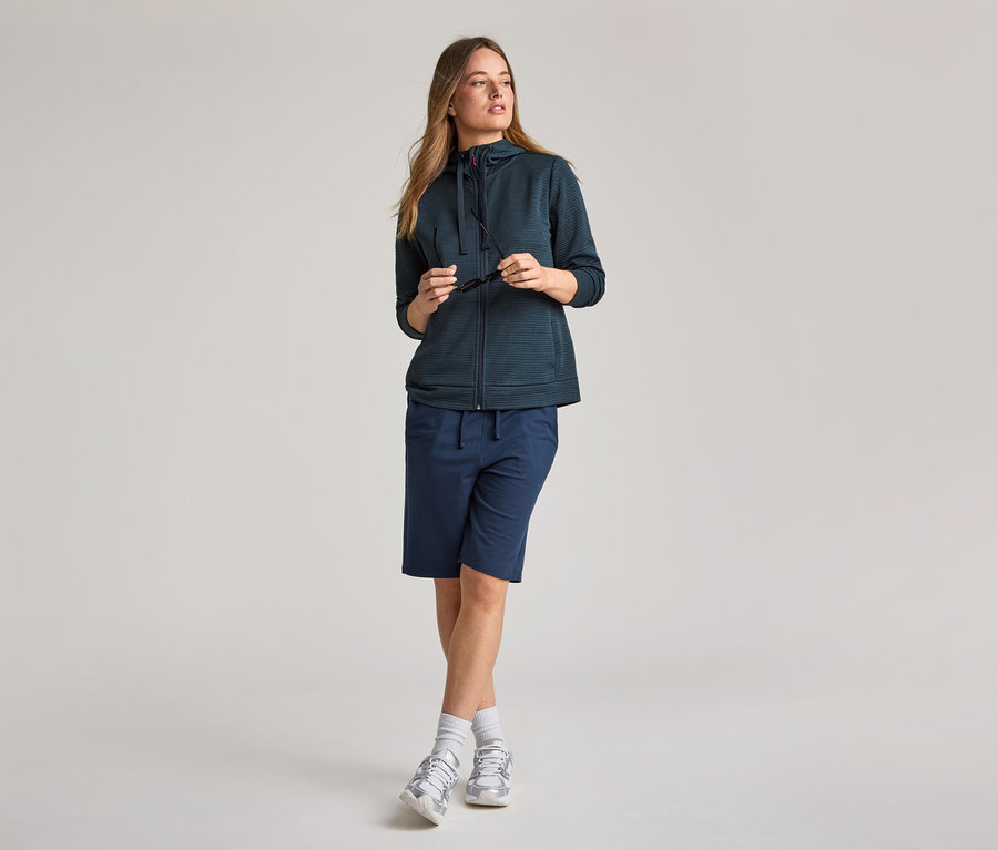 Žena v námorníckej modrej mikine s kapucňou a šortkách. Drží slnečné okuliare.