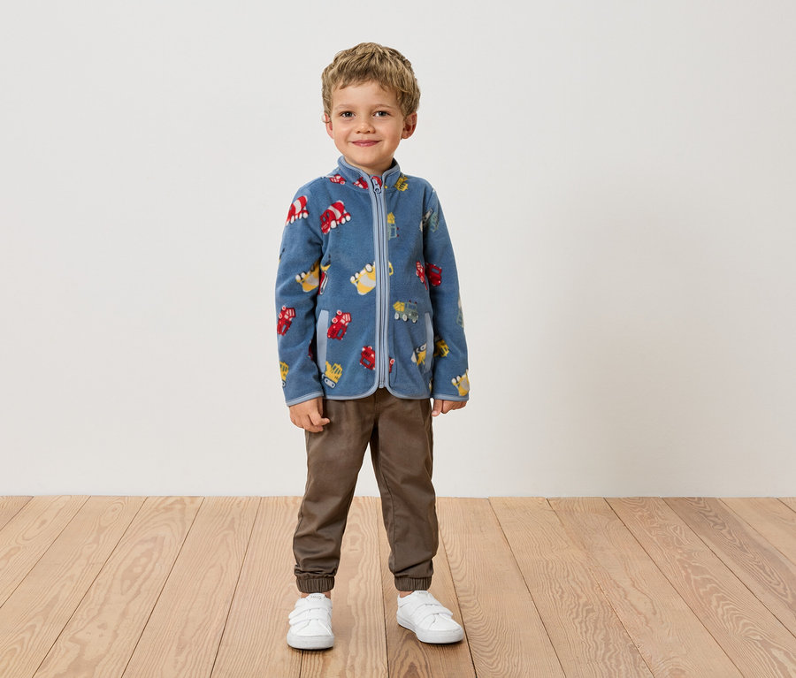 Chlapec stojí pred stenou v modrej fleecovej bunde so vzorom vozidiel, hnedých nohaviciach a bielych topánkach.