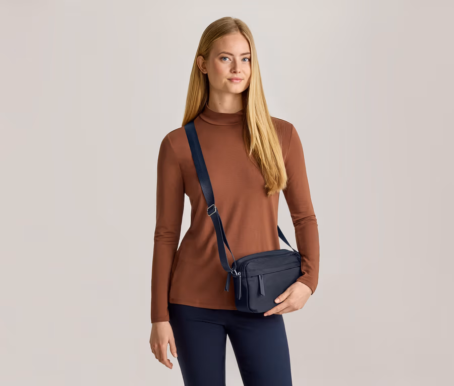 Žena s dlhými blond vlasmi má na sebe hnedý top s dlhým rukávom, tmavomodré nohavice a tmavomodrú crossbody tašku. Tašku drží ľavou rukou a pozerá sa dopredu.