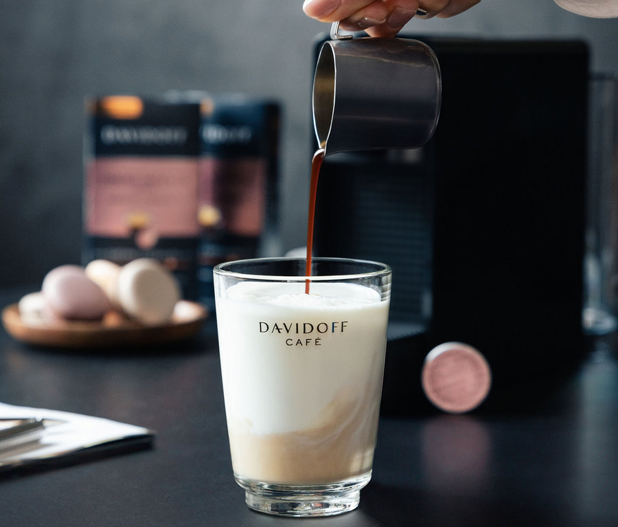 Ruka nalieva kávu do pohára s mliekom. V pozadí je kávovar a balenie DAVIDOFF CAFÉ Crema Intense.