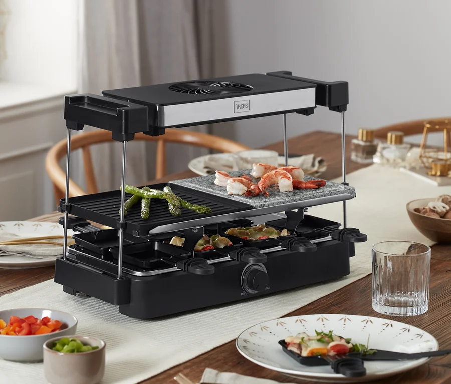 Na drevenom stole stojí raclette gril Trebs s digestorom a kamennou doskou »15100«, na ktorom sa grilujú jedlá.