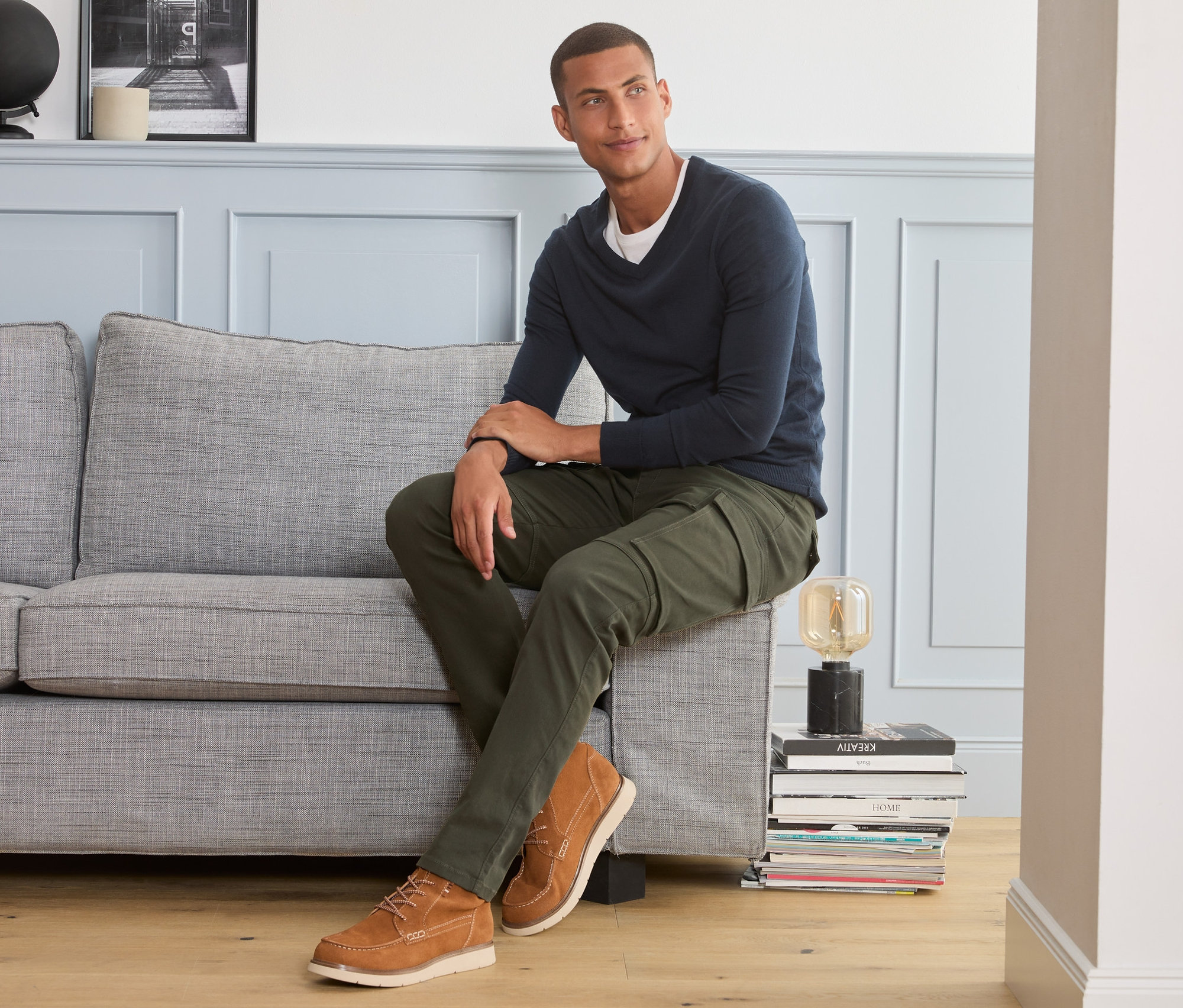 Muž sedí na sivej pohovke a má na sebe cargo nohavice.