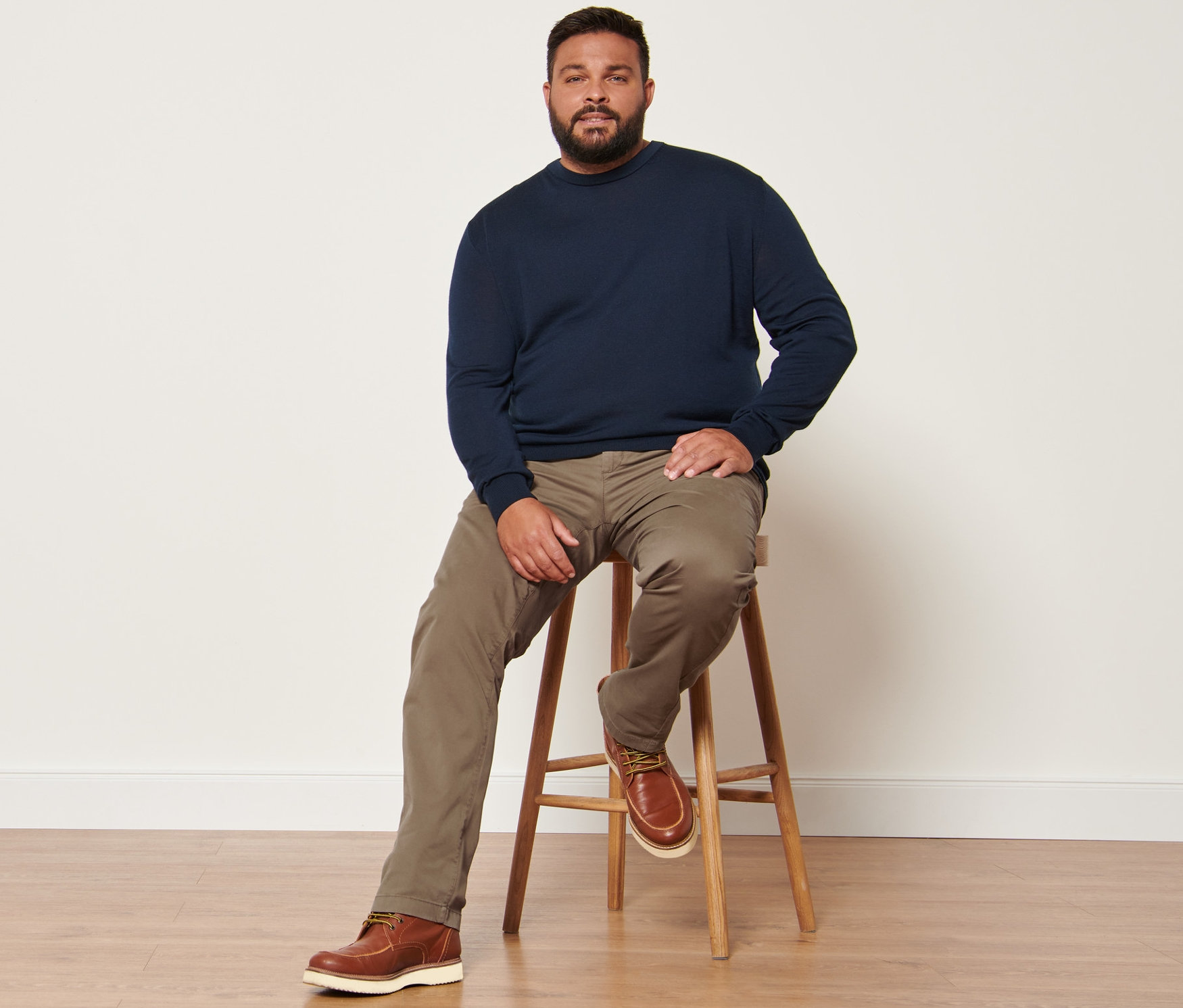 Muž sedí na stoličke a má na sebe námornícky modrý pulóver z vlny merino a pieskové nohavice chino.