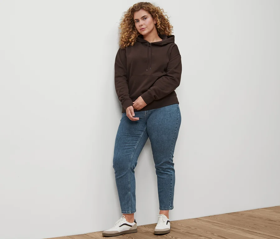 Žena stojí a má na sebe rovné džínsy »Fit Juna« stredne modré, kožené topánky sneaker a tmavohnedú mikinu s kapucňou.