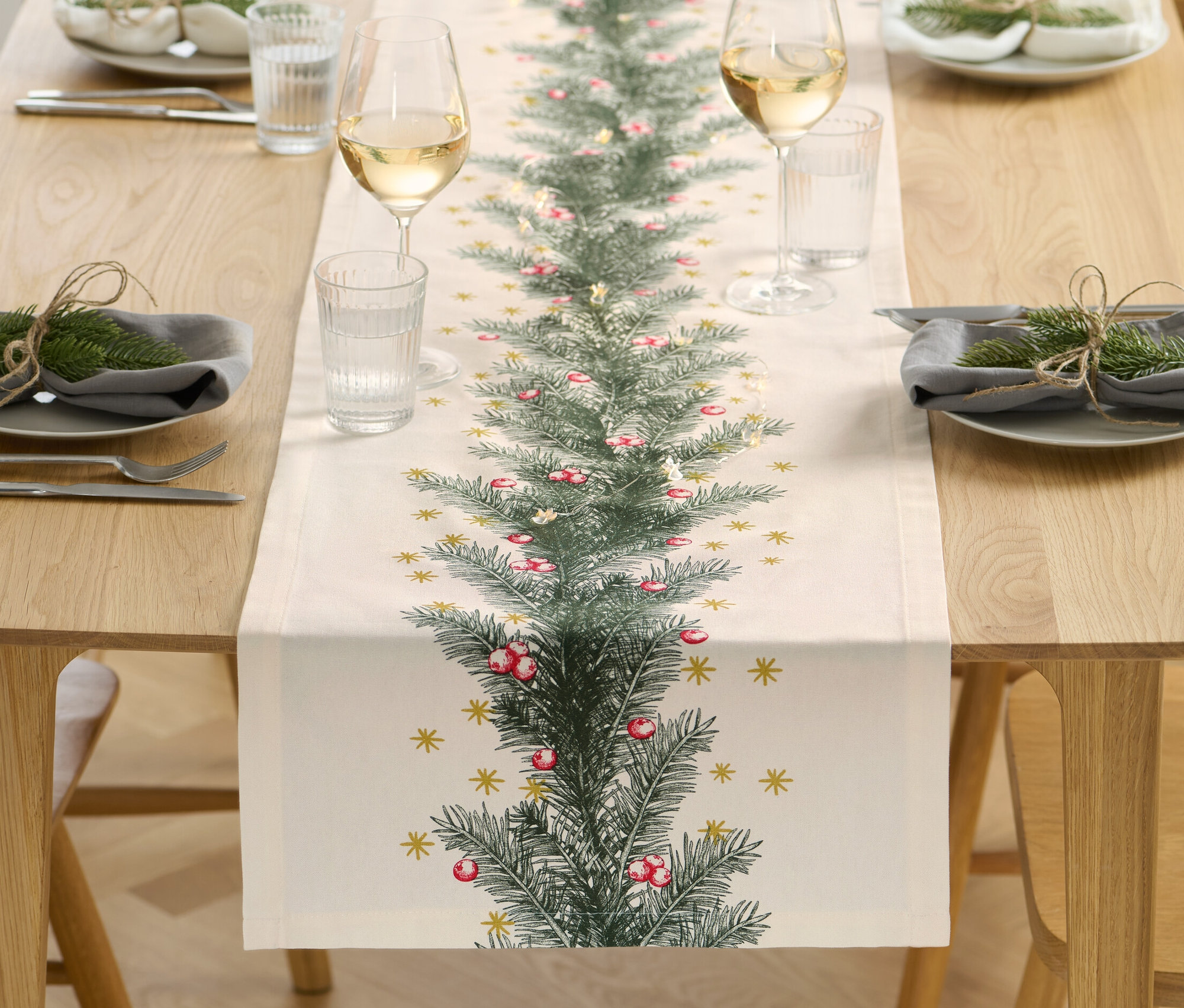 Uprostred dreveného stola leží behúň s vianočnou potlačou. Po oboch stranách sú prestreté taniere, poháre a príbory.