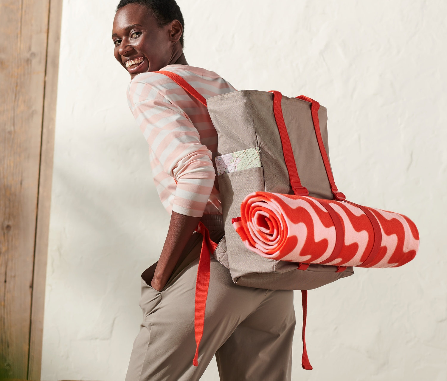 Usmievajúca sa žena s piknikovým ruksakom s červenými popruhmi a zrolovanou dekou s červeno-bielym vzorom.