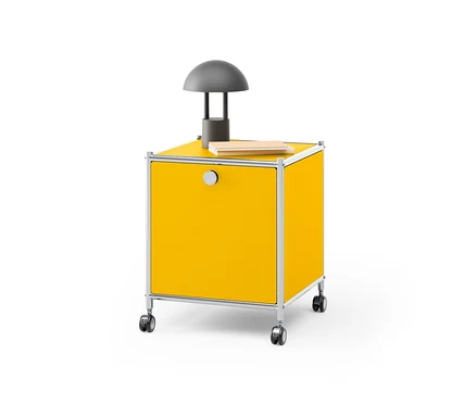 Žltý kovový nočný stolík »CN3« s výklopným priečinkom, na ktorom stojí sivá lampa a leží kniha.