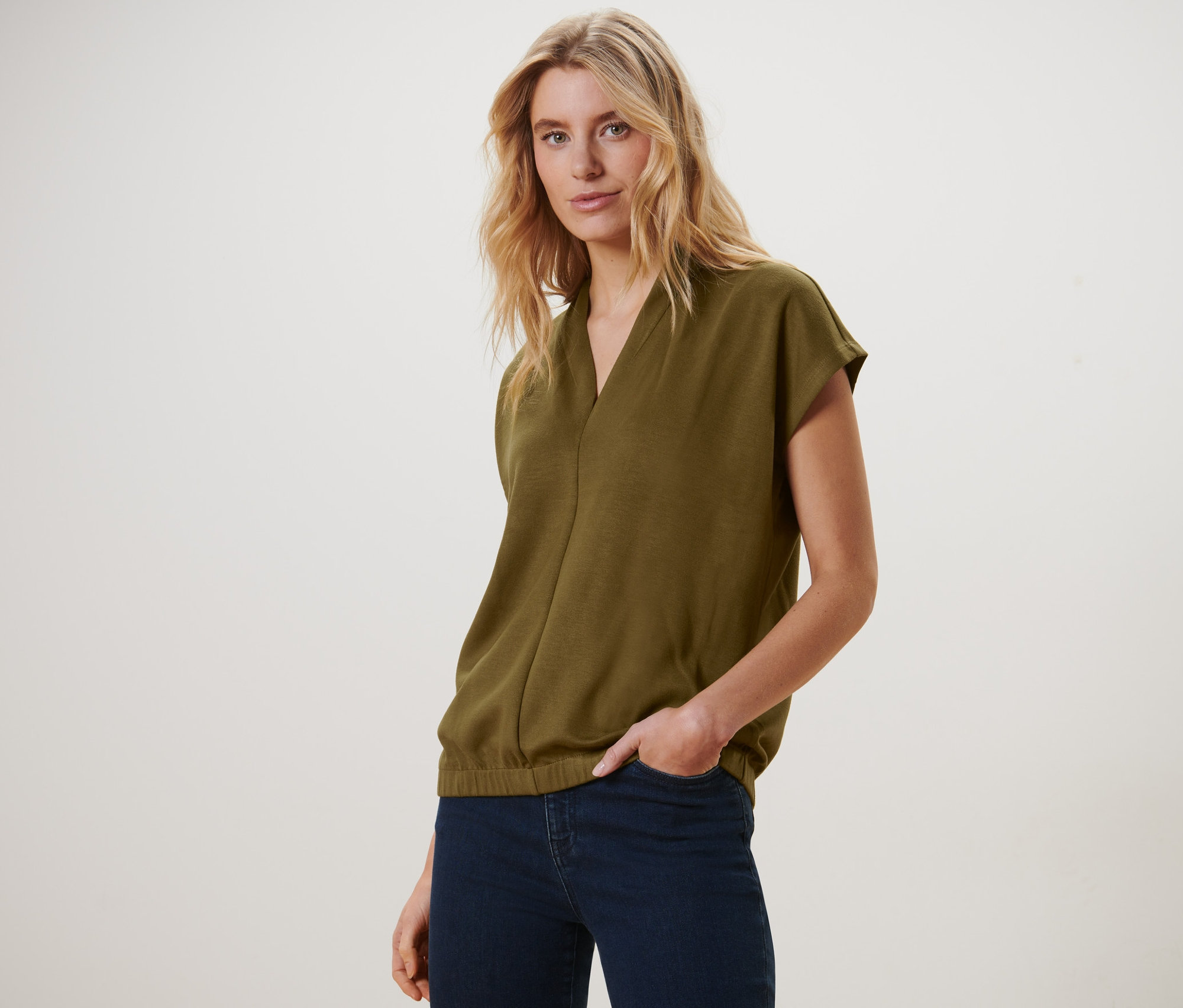 Blondínka pózuje v olivovozelenom blúzkovom tričku s výstrihom do V a tmavomodrých džínsoch.