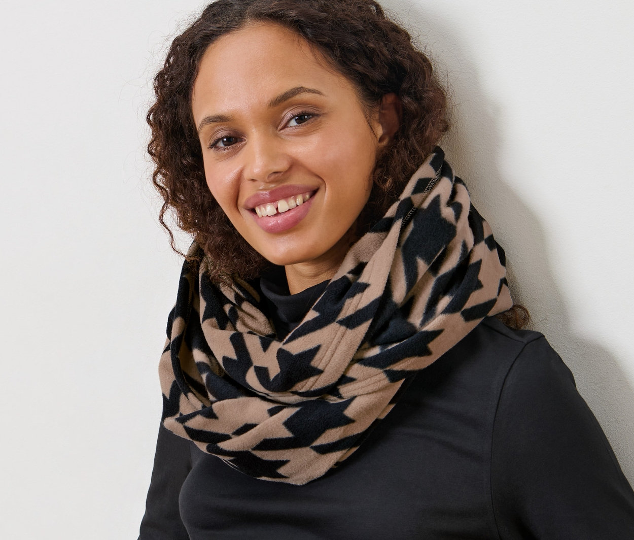 Žena s hnedými kučeravými vlasmi sedí a má na sebe flaušový kruhový šál s potlačou kohútej stopy.
