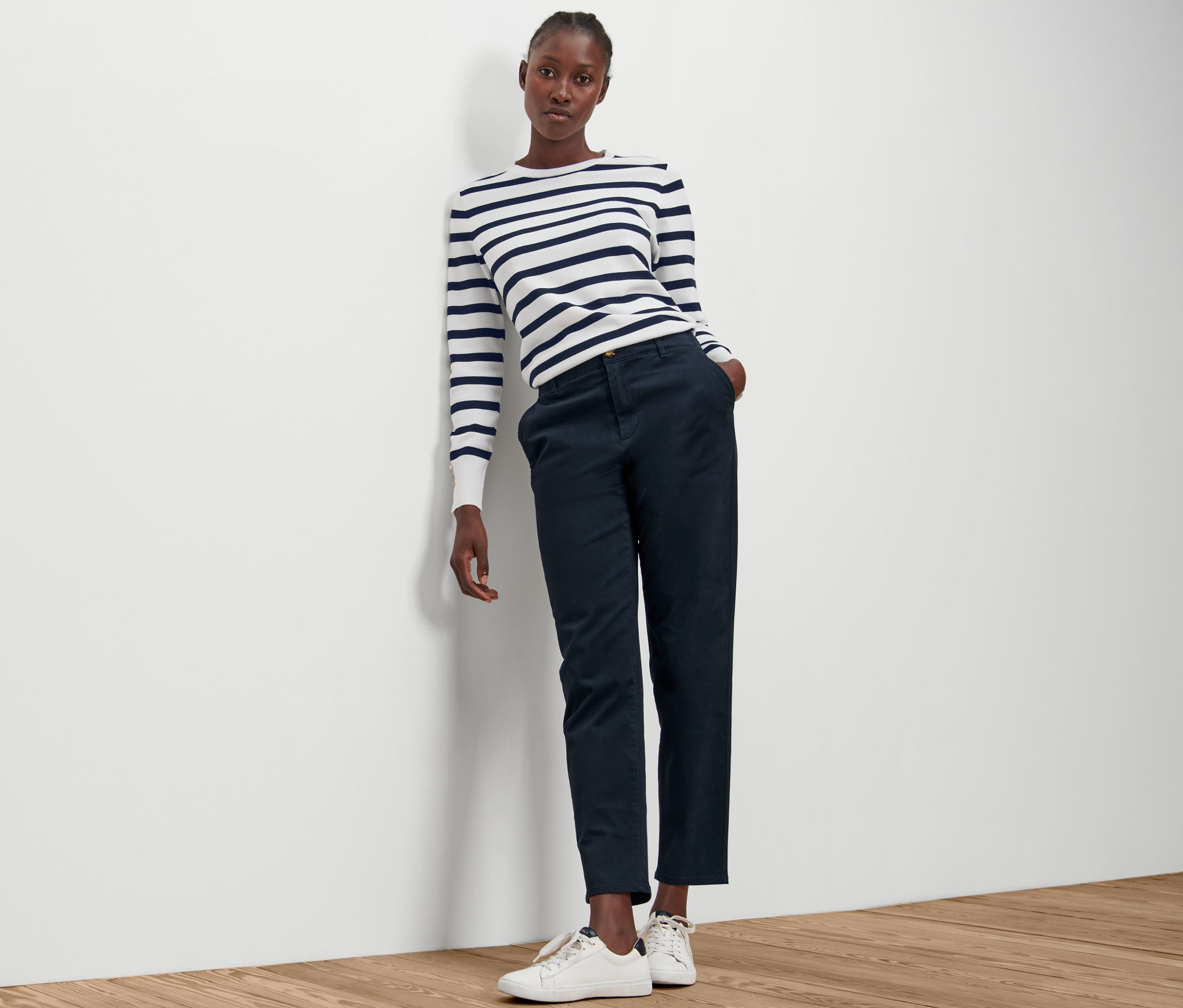 Žena opretá o stenu má na sebe pruhovaný top a námornícke modré chino nohavice.