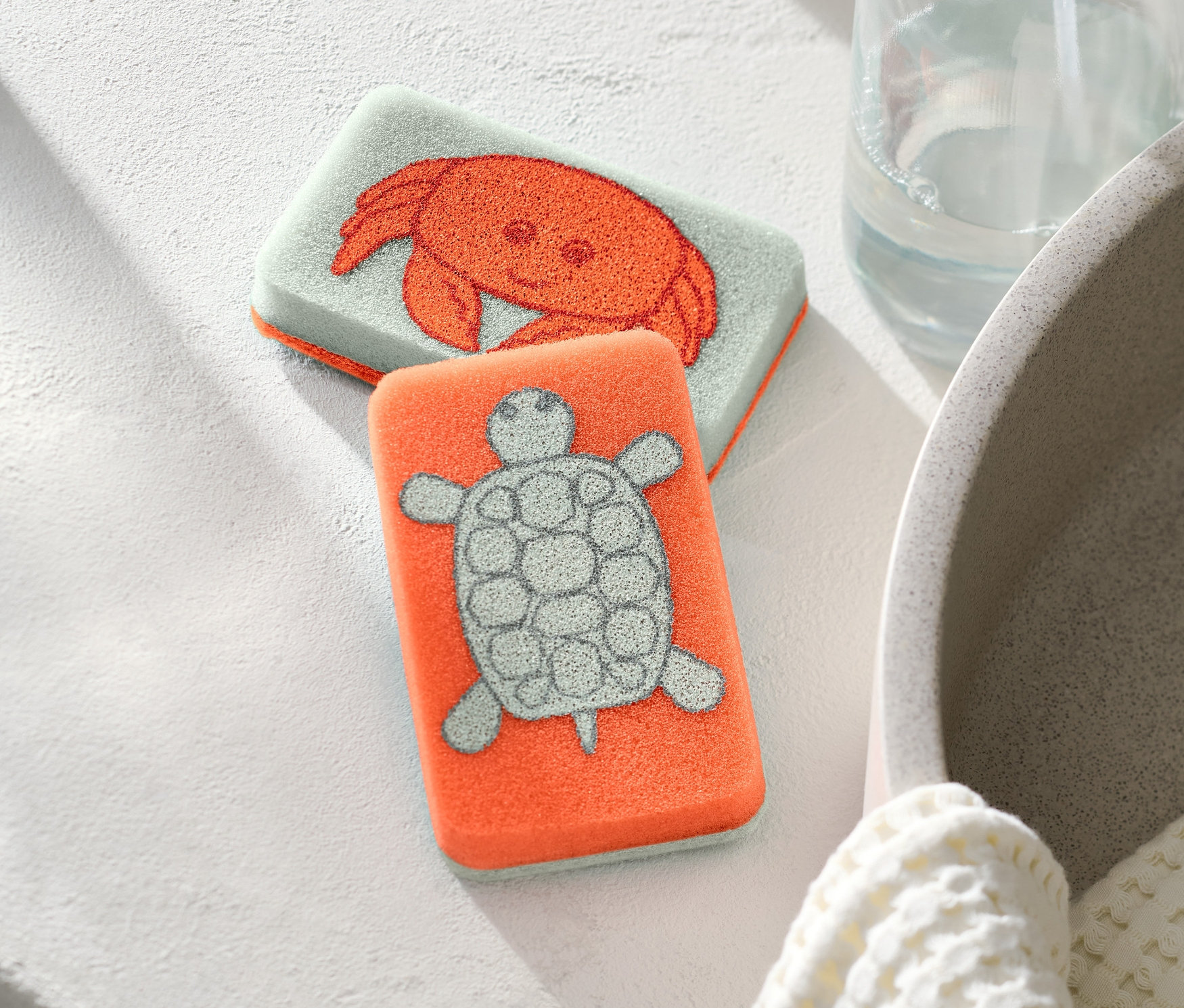 Dve čistiace špongie, jedna s motívom kraba a druhá s motívom korytnačky, ležia vedľa seba.