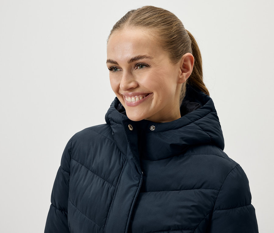 Žena s hnedými vlasmi stiahnutými do chvosta sa usmieva a má na sebe tmavomodrú páperovú bundu.