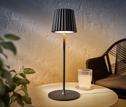 Čierna LED akumulátorová stolová lampa stojí na stole vedľa pohára s nápojom.