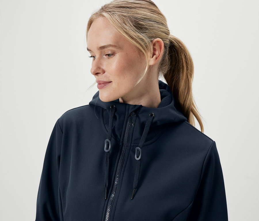 Portrét ženy s blond vlasmi stiahnutými do copu, oblečenej v tmavomodrej mikine s kapucňou a zipsom.